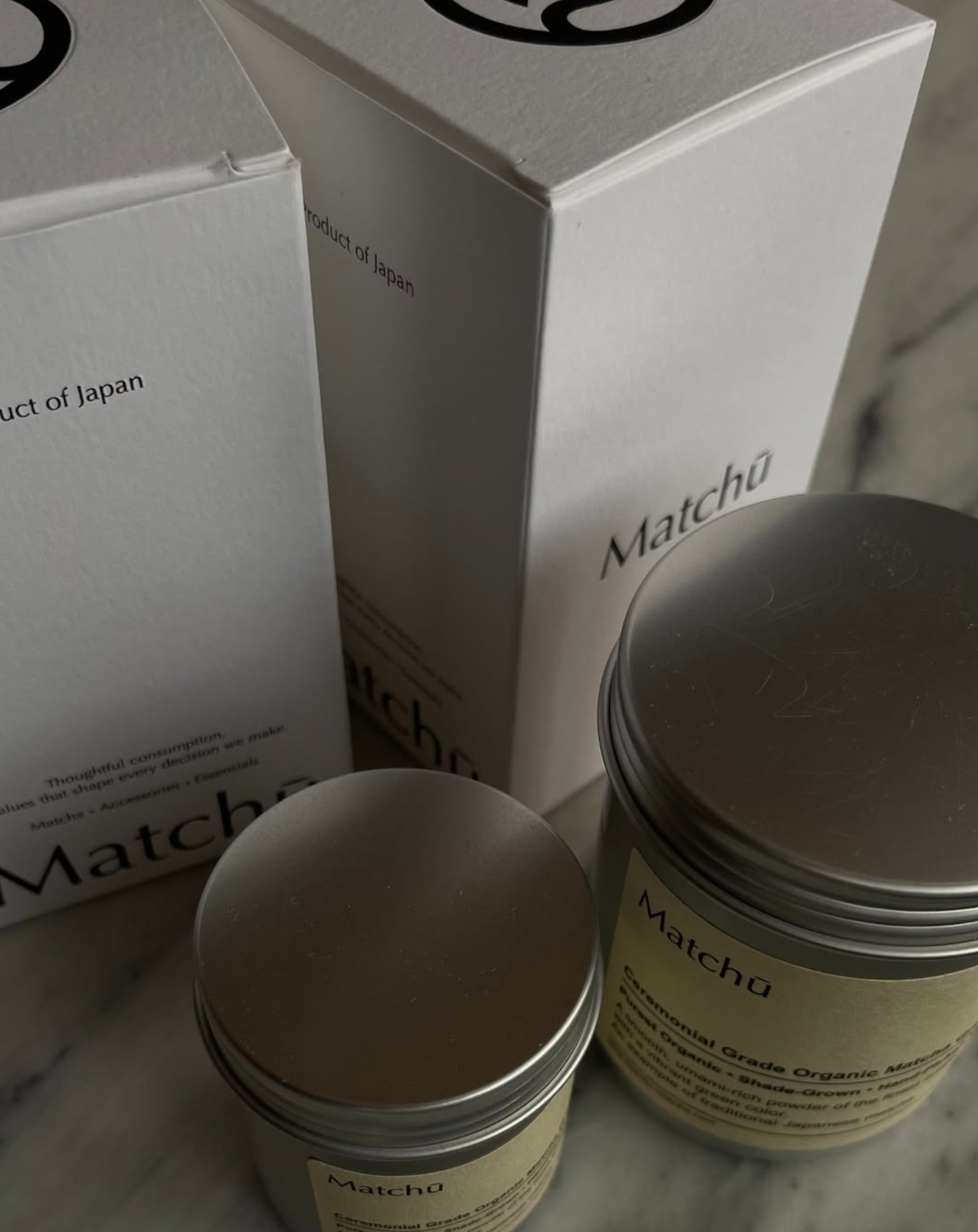 Two Matchu Matcha containers on a marble surface with boxes in the background.