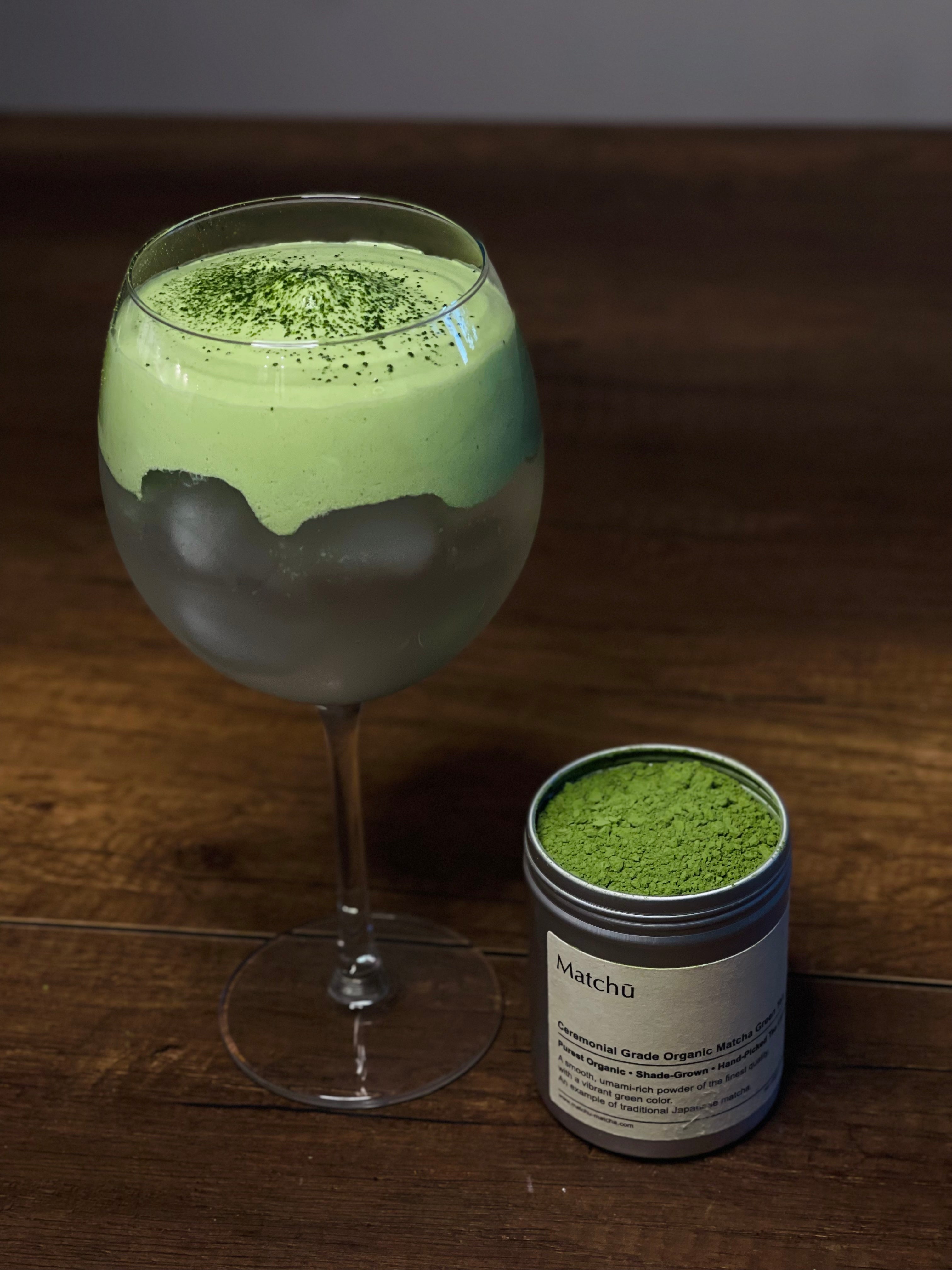 Matchu Matcha green tea latte in a glass with a container of matcha powder on a wooden surface.